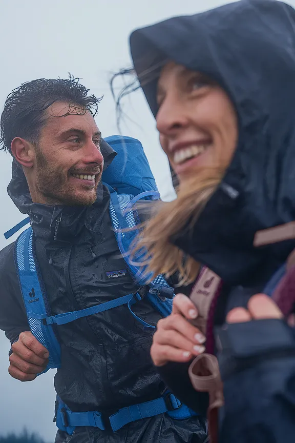 Zwei lächelnde Personen in Outdoor-Kleidung bei Regen, einer trägt einen blauen Rucksack.