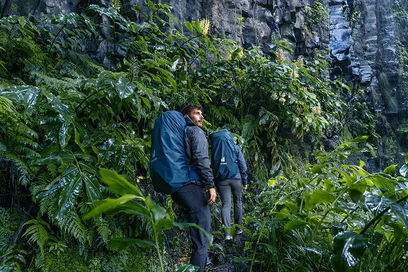 Wanderer mit blauem Wanderrucksack auf nassem Dschungelpfad an einer Felswand.