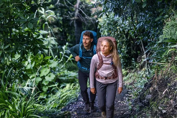 Zwei Wanderer mit Wanderrucksäcken auf einem steilen, von Farnen gesäumten Waldpfad.